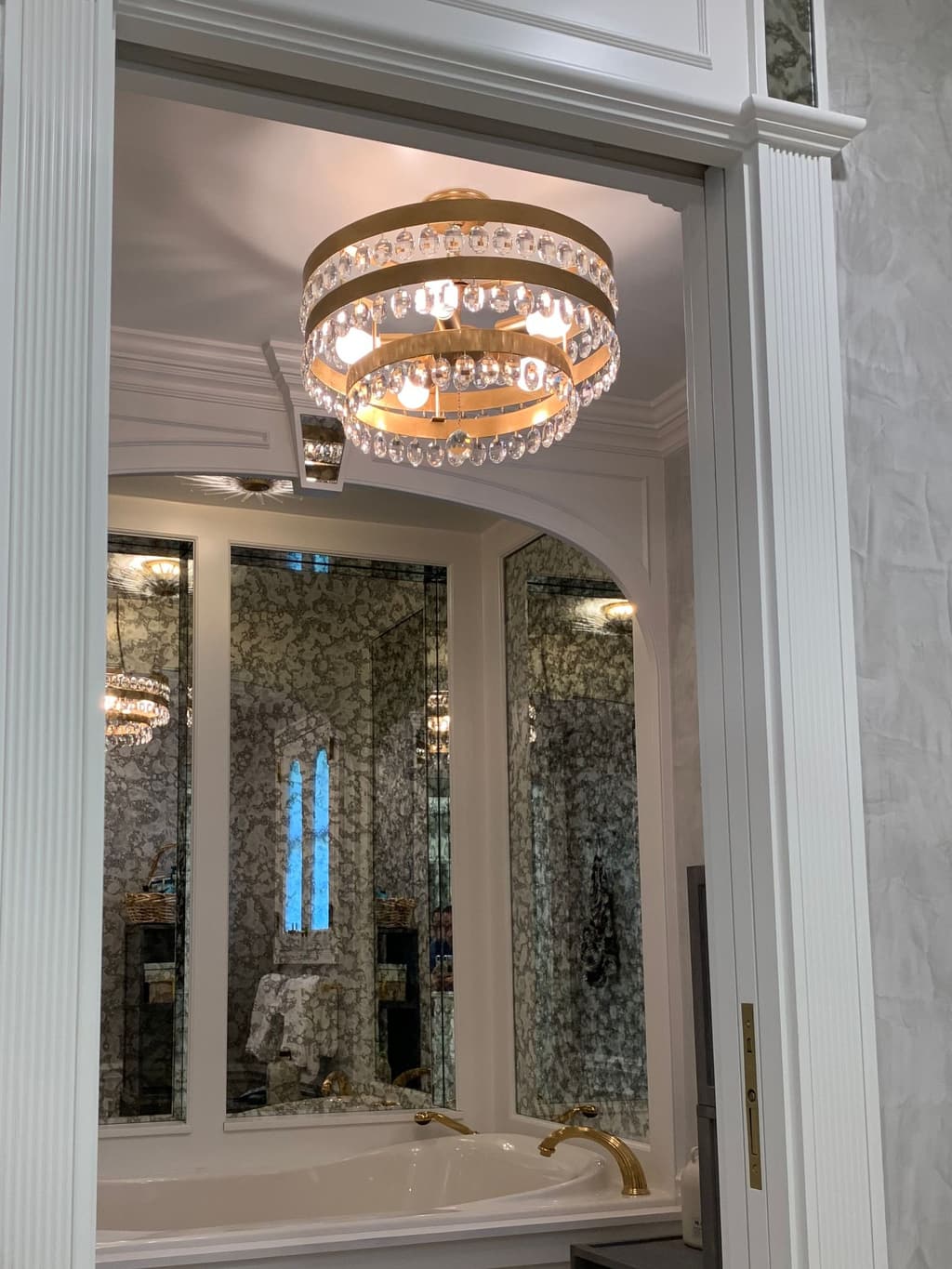 A tub alcove framed by fluted white pilasters, with a crystal and gold chandelier, mirrored panels and softly mottled plaster walls.