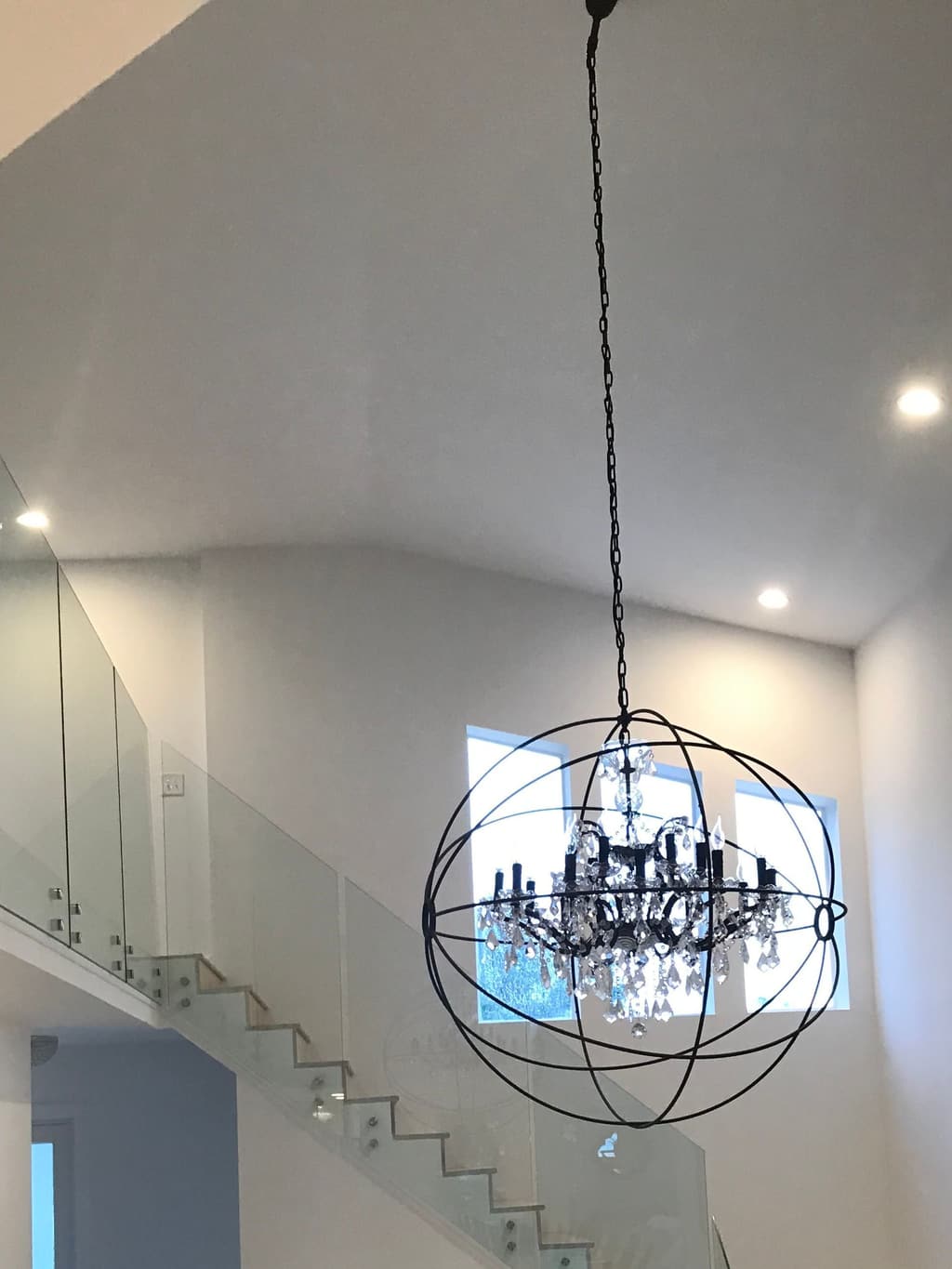 The finished stair hall: a frameless glass rail runs up the open stair and along the landing, under the orb chandelier.